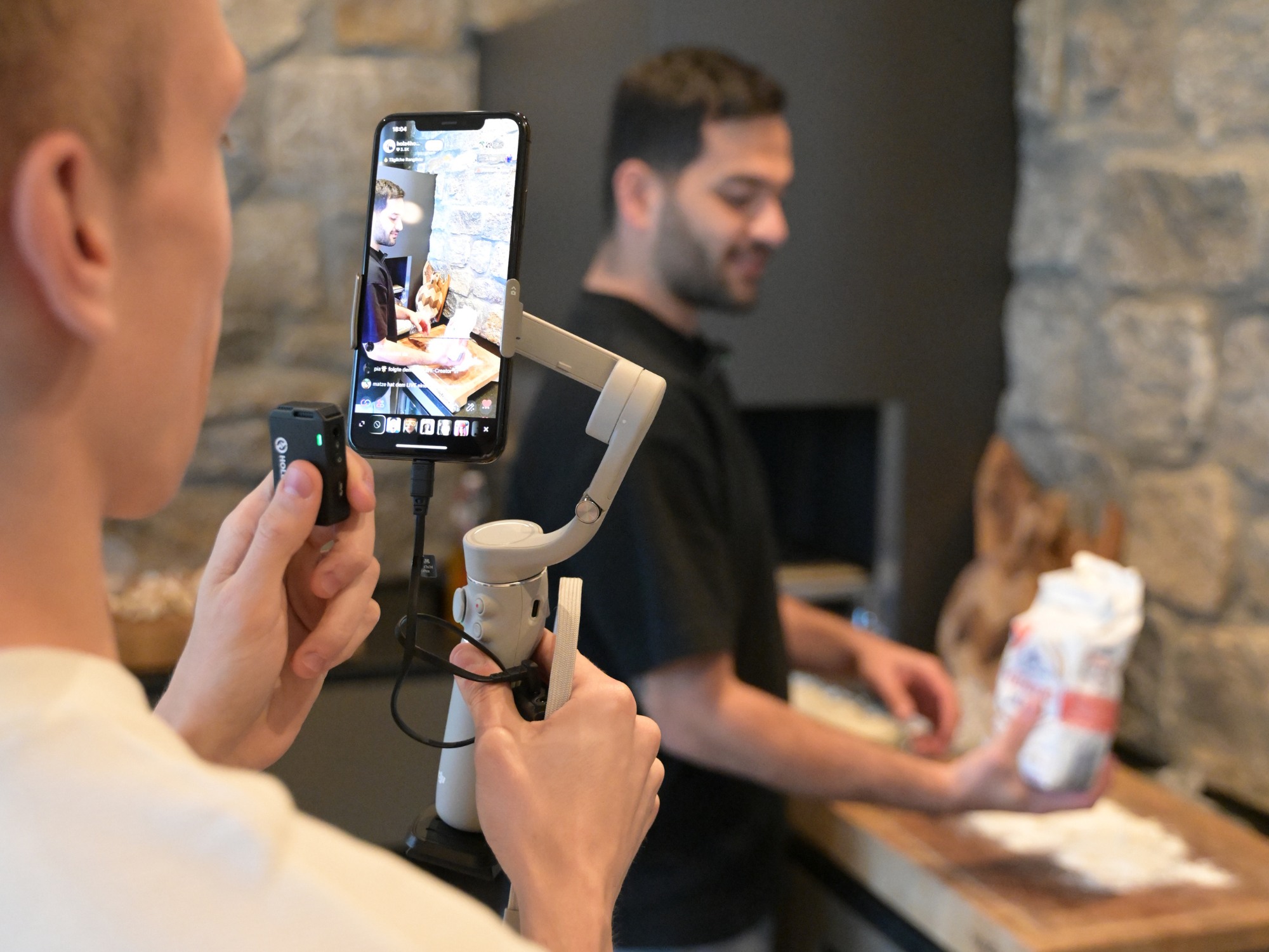 Zwei Männer produzieren Content mit Smartphone und Gimbal vor einer Steinwand am Kamin