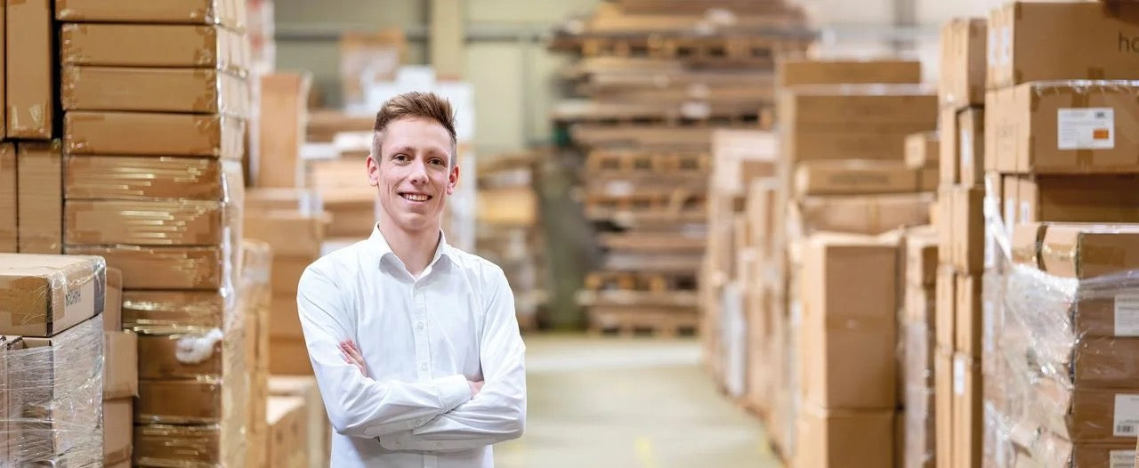 Junger Mann im weißen Hemd steht mit verschränkten Armen in einem Lager voller verpackter Möbel